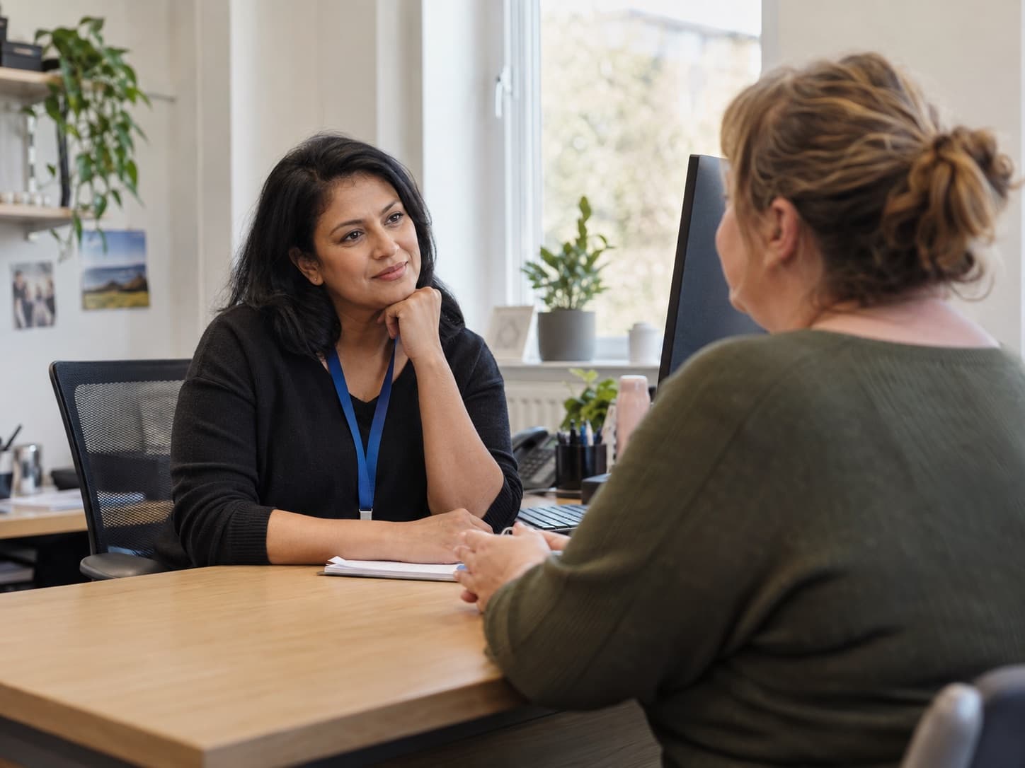 A prescriber listening to a patient during a medical weight management consultation, across a desk in a daylit consulting room